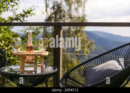 Champagner- und Cocktailgläser auf dem Glastisch auf der Außenterrasse mit Blick auf die Berge bei Sonnenuntergang Stockfoto