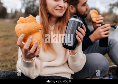 Das Paar trinkt aus einem Thermalbecher und isst Croissants auf einem Spaziergang im Park. Stockfoto