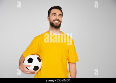 Glücklicher bärtiger Fußballfan, der den Fußball hält und isoliert auf grauem, stockem Bild wegschaut Stockfoto
