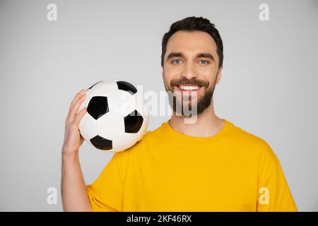 Zufrieden bärtiger Fußballfan in gelbem T-Shirt, der den Fußballball hält und die Kamera isoliert auf grauem, Stock-Bild anschaut Stockfoto
