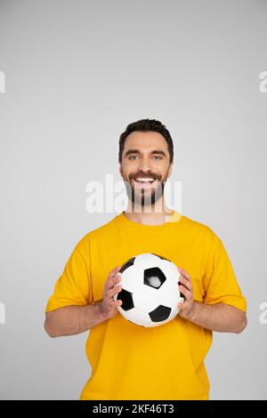Fröhlicher bärtiger Sportfan mit Fußballball, der die Kamera isoliert auf einem grauen Bild betrachtet Stockfoto