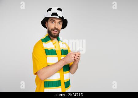 Positiver und neugieriger Mann in Fußballfanhut und gestreiftem Schal, der Smartphone hält und die Kamera isoliert auf grauem, stockem Bild anschaut Stockfoto