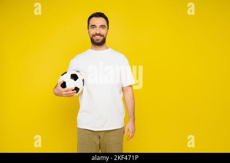 Glücklicher bärtiger Mann in weißem T-Shirt, der mit Fußballball steht und die Kamera isoliert auf gelbem, Stockbild anschaut Stockfoto
