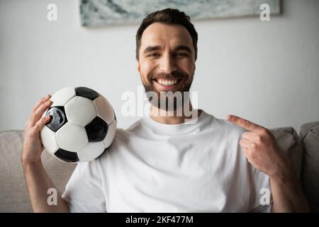 Glücklicher bärtiger Mann, der die Kamera ansah und zu Hause auf den Fußball zeigte, Stockbild Stockfoto