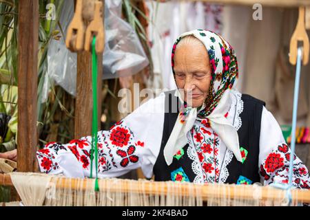 Eine alte weißrussische oder ukrainische Frau in einem gestickten Hemd an einem Vintage-Webstuhl. Slawische ältere Frau in ethnischer Kleidung. Stockfoto