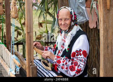 Eine alte weißrussische oder ukrainische Frau in einem gestickten Hemd an einem Vintage-Webstuhl. Slawische ältere Frau in ethnischer Kleidung. Stockfoto