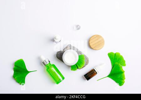 Frische grüne Ginkgo-Blätter, kosmetische Serumflasche und Cremedose auf weißem Hintergrund. Natürliche handgemachte Kosmetik-Konzept. Stockfoto