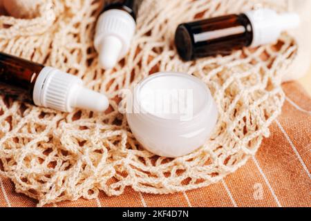 Cremefarbenes Glas auf einem Netzbeutel und braune Glasflaschen mit ätherischen Ölen auf orangefarbenem Stoffhintergrund. Bio Spa kosmetisches Beauty-Produkt. Keine Abfallentsorgung Stockfoto