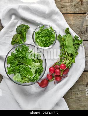 Frischer grüner Salat in Schüsseln, Brokkoli und Radieschen auf weißem Textilhintergrund Stockfoto