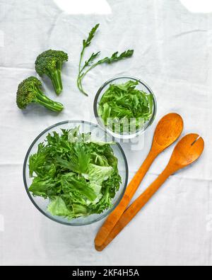 Frischer grüner Salat in Glasschüsseln und Brokkolistücke mit Holzlöffeln auf weißem Hintergrund Stockfoto