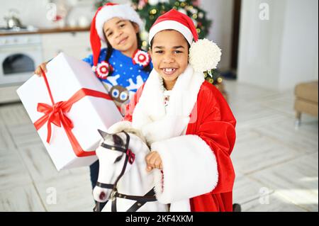 Ein Bruder und eine Schwester im Weihnachtsmann-Kostüm und ein Pullover mit einem Hirsch reiten auf einem schaukelnden Pferd und halten eine riesige Schachtel mit einem Weihnachtsgeschenk in den Händen Stockfoto