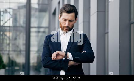 Ein professioneller, selbstbewusster Geschäftsmann, der draußen auf ein Treffen wartet, sieht auf die Armbanduhr, die die Zeit überprüft, unzufrieden kaukasischer Mann, der auf die Uhr zeigt Stockfoto