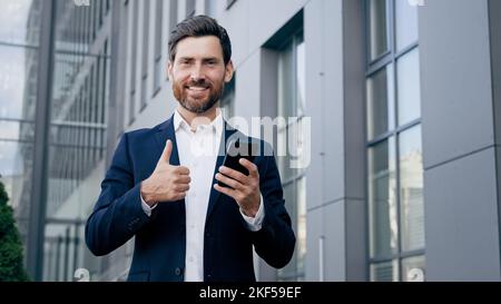 Glücklicher Mann, der im Freien steht und ein lächelndes Telefon hält, Geschäftsmann, der die neue mobile App für geschäftliche Aufgaben verwendet und dabei den Daumen nach oben zeigt Stockfoto