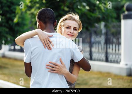 Porträt eines jungen afroamerikanischen Mannes, der mit dem Rücken im Freien steht und zum Dank eine attraktive Frau mittleren Alters umarmt. Zuneigung, Abschied Stockfoto