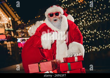 Foto von funky aufgeregt erstaunt santa Carry präsentiert Tasche Stapel geschenkbox Girlande Dekor Neujahr Baum im Freien Stockfoto