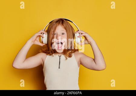 Schreiende kleine stilvolle Mädchen mit langen roten Haaren und Sommersprossen hören Musik isoliert über hellgelben Hintergrund. Konzept der Kinder Emotionen Stockfoto