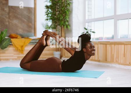 Schöne und athletische afro amerikanische Frau, die mit einem Lächeln in der Bogenstellung Seitenansicht liegt und Yoga auf einer Gymnastikmatte im dekorierten Studio macht. Muskeldehnung Stockfoto