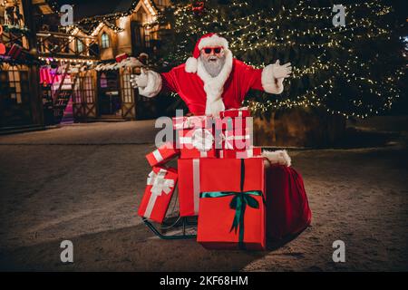 Foto in voller Größe von verrückt überglücklich Großvater santa gratulieren Neujahr Stapel geschenkbox Baum Lichter Girlande Innenstadt im Freien Stockfoto