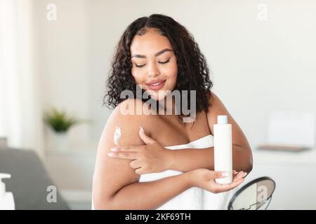 Schöne afroamerikanische mollige Frau, die nach dem Bad in ein Handtuch gehüllt wurde und Körpercreme auf ihre Schulter aufbrachte und im Schlafzimmer saß Stockfoto