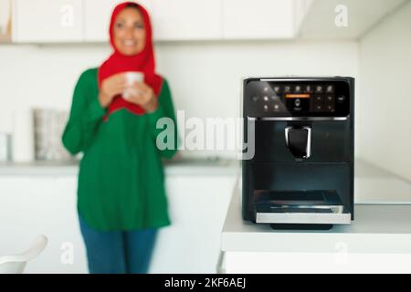 Aufgeregte muslimische Dame mittleren Alters trinkt morgendlichen Kaffee in der Küche, konzentrieren Sie sich auf moderne Kaffeemaschine Stockfoto