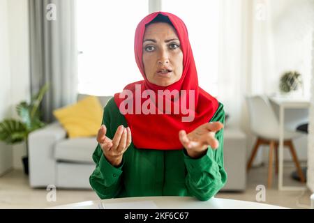 Reife islamische Frau, die Hijab trägt und mit der Kamera spricht, online kommuniziert, im Wohnzimmer sitzt Stockfoto