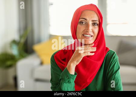 Glückliche muslimische Frau mittleren Alters, die roten Hijab trägt, vor der Kamera posiert und lächelt und zu Hause im Wohnzimmer sitzt Stockfoto