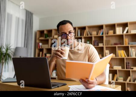 Arabischer Mann, der von zu Hause aus online studiert, Notebook liest und Kaffee trinkt, mit Laptop am Schreibtisch sitzt, freier Platz Stockfoto