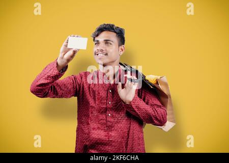 Junger, glücklicher Inder, der Einkaufstasche und leere Karte hält, während er traditionelle Tücher trägt, die vor gelbem Hintergrund stehen Stockfoto