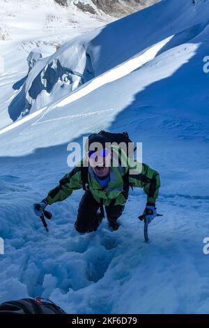 Beschreibung: Begeisterter Bergsteiger klettert mit Eisachsen über einen steilen Schneehang entlang der Jungfrau-Höhenroute mit Aletschgletscher im Backgro Stockfoto