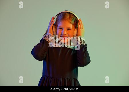 Musik. Schöne rothaarige kleine Mädchen trägt festliches Kleid Blick auf Kamera isoliert auf blauem Hintergrund in Neonlicht. Kinder Emotionen, Gesichtsbehandlung Stockfoto