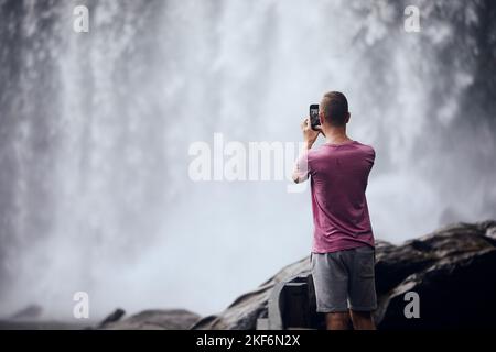 Rückansicht des Touristen beim Fotografieren mit dem Handy. Mann vor hohem Wasserfall in Kambodscha. Stockfoto