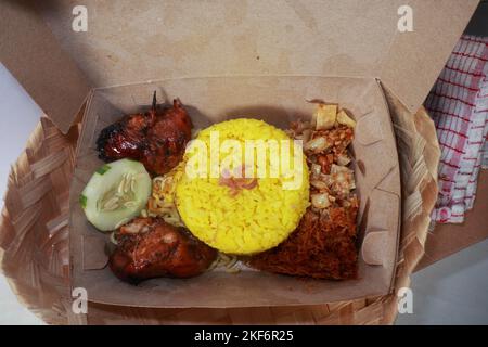 Draufsicht auf traditionelle indonesische Küche, nämlich Nasi Kuning mit zwei gegrillten Hühnern, gebratener Kokosraspel, einfachen Nudeln, getrocknetem Tempeh und einer Scheibe Stockfoto