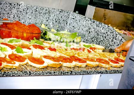 Salat- und Tomatenplatten auf der Arbeitsplatte, Burger nacheinander mit pommes frites und Tomatenscheiben geöffnet. Herd-Hand hält eine küchenzange und füllt sie Stockfoto