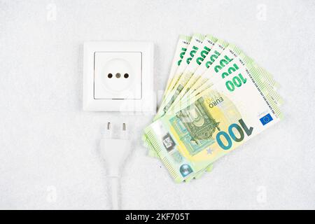 Weiße Steckdose mit Stecker mit vielen 100-Euro-Banknoten auf hellem Hintergrund. Konzept der steigenden Strompreise in Europa. Hohe Energiekosten, w Stockfoto