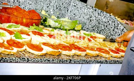Salat- und Tomatenplatten auf der Arbeitsplatte, Burger nacheinander mit pommes frites und Tomatenscheiben geöffnet. Herd-Hand hält eine küchenzange und füllt sie. Stockfoto