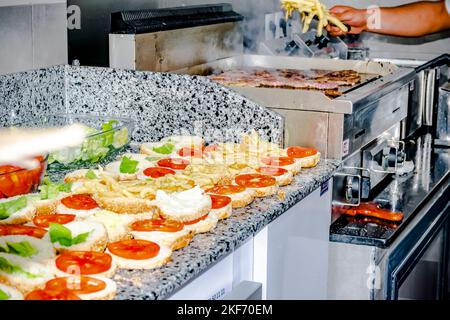 Offene Burger auf der Arbeitsplatte mit pommes Frites, Salat und Tomate. Herd hält eine küchenzange, um die Hamburger zu füllen. Fleisch wird auf dem Grill gekocht. Stockfoto