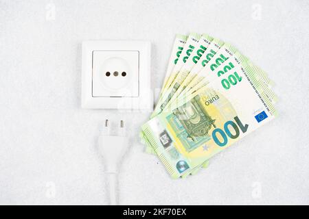 Weiße Steckdose mit Stecker mit vielen 100-Euro-Banknoten auf hellem Hintergrund. Konzept der steigenden Strompreise in Europa. Hohe Energiekosten, w Stockfoto