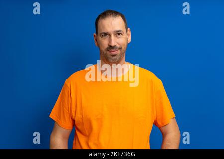 Isolierte Aufnahme eines gutaussehenden bärtigen Mannes, trägt ein lässiges orangefarbenes T-Shirt, hat einen fröhlichen Ausdruck, während er seinem Gesprächspartner zuhört, posiert im Studio Stockfoto