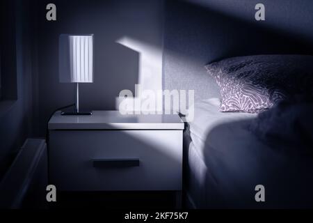 Mondlicht im Schlafzimmer in der Nacht vom Fenster. Bett und Tisch in dunklem Raum in der Abenddämmerung. Innenarchitektur in modernen weißen Apartment. Blaues Licht vom Mond. Stockfoto