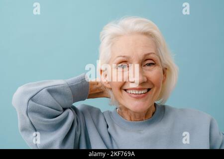 Schöne glückliche ältere Frau in blauem Pullover, die ihr Haar auf einem isolierten blauen Hintergrund anpasst Stockfoto