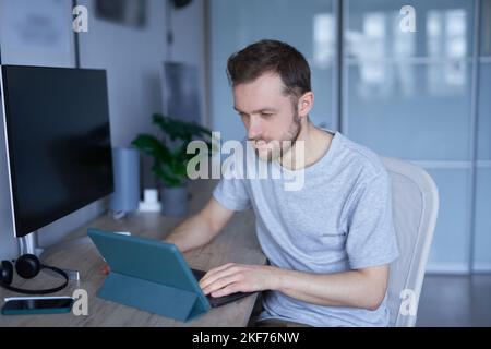 Attraktiver bärtiger Mann, der im Heimbüro mit einem Tablet sitzt, das online arbeitet oder unterrichtet. Nahaufnahme des Porträts eines jungen, hübschen Mannes in grauem Hemd. Hochwertige Fotos Stockfoto