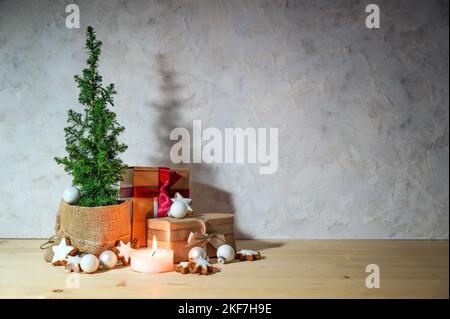 Alternativer Weihnachtsbaum, kleine Topfkoniferpflanze, geschmückt mit Sternen, Kugeln, Geschenken und einer Kerze auf einem hellen Holztisch gegen einen rus Stockfoto