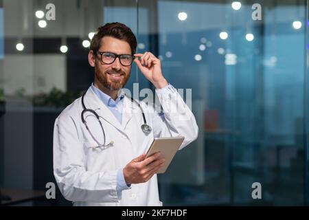Porträt eines jungen hübschen männlichen Arztes. Er steht im Büro in einem weißen Mantel und mit einem Stethoskop. Er hält eine Tablette in den Händen. Er schaut auf die Kamera, passt seine Brille an, lächelt. Stockfoto
