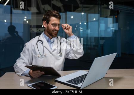 Ein junger Arzt spricht mit einem Patienten über einen Videoanruf von einem Laptop. Er sitzt an einem Tisch in einem Büro in einem Krankenhaus. Er hält einen Ordner mit Dokumenten, passt seine Brille an, spricht, lächelt. Stockfoto