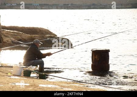 Sliema, Malta - 12. November 2022: Fischer sitzen auf einem Stuhl auf den Felsen am Meer, während sie an einem sonnigen Morgen mit Fischerstick fischen Stockfoto