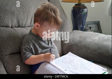 Kleiner Junge, der zu Hause auf einer Couch sitzt und in einem Notizbuch Kritzeleien zeichnet Stockfoto