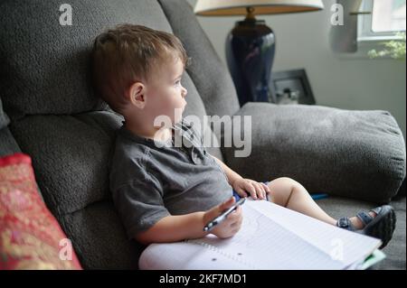 Kleiner Junge, der zu Hause auf einer Couch sitzt und in einem Notizbuch Kritzeleien zeichnet Stockfoto