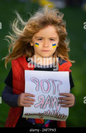 Kind Junge hält Schild mit Worten Stoppt den Krieg im Freien stehen. Kind trägt ein Schild Krieg stoppen. Putin greift das ukrainische Volk an. Stockfoto