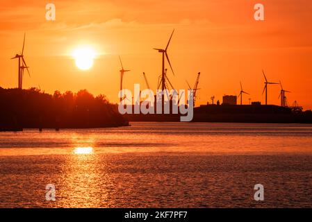 Industriegebäude, Windturbinen und Dockkrane an einem Flusshafen, die bei Sonnenuntergang vor einem feurigen Himmel geschildet wurden Stockfoto