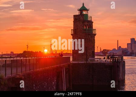 Steinlichtturm an einem Flusshafen unter einem dramatischen Himmel bei Sonnenuntergang. Die Leute, die den Sonnenuntergang bewundern, sind auf dem Pier. Industriegebäude befinden sich im Hintergrund. Stockfoto